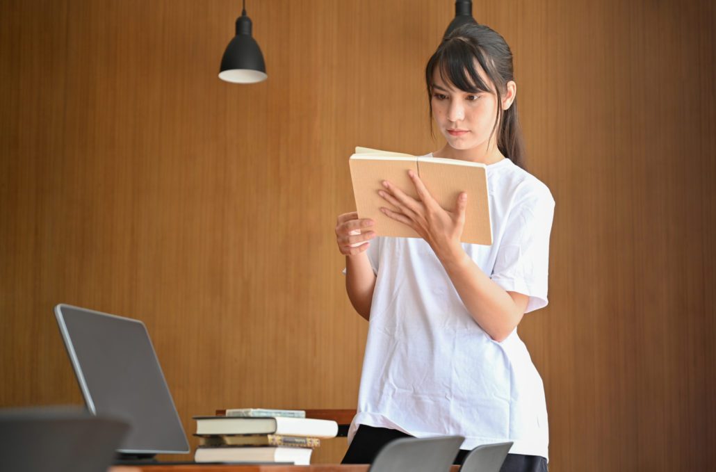 Young female student standing in library and reading a book.